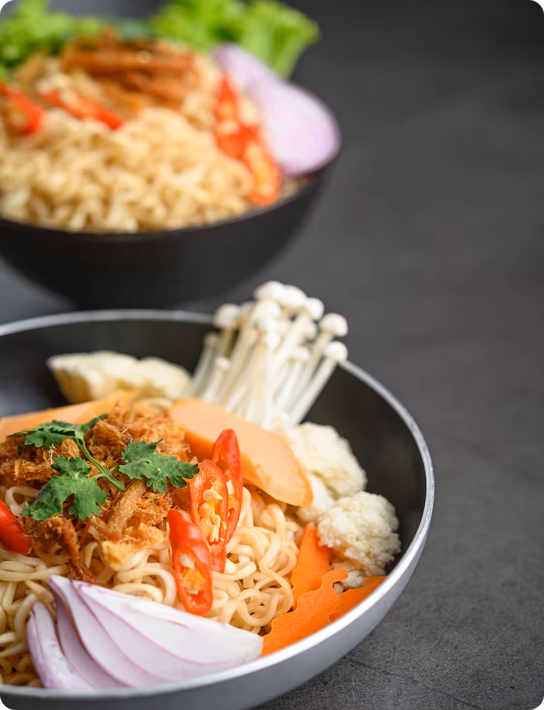 Bowl of noodles garnished with sliced red chili peppers, cilantro, fried shallots, red onion slices, enoki mushrooms, carrot slices, and cauliflower.